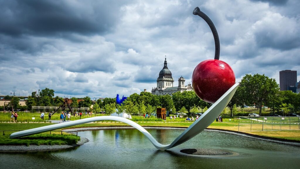 Photograph of Spoonbridge and Cherry, a sculpture and fountain shaped like a giant bent silver spoon with a red cherry balanced at the tip. 